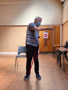 An older white man in a light hooped t-shirt and facemask does an exercise using red resistance bands