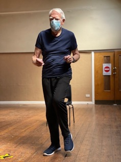 An older white man in a navy T-shirt and facemask walks from a chair as part of an axercise